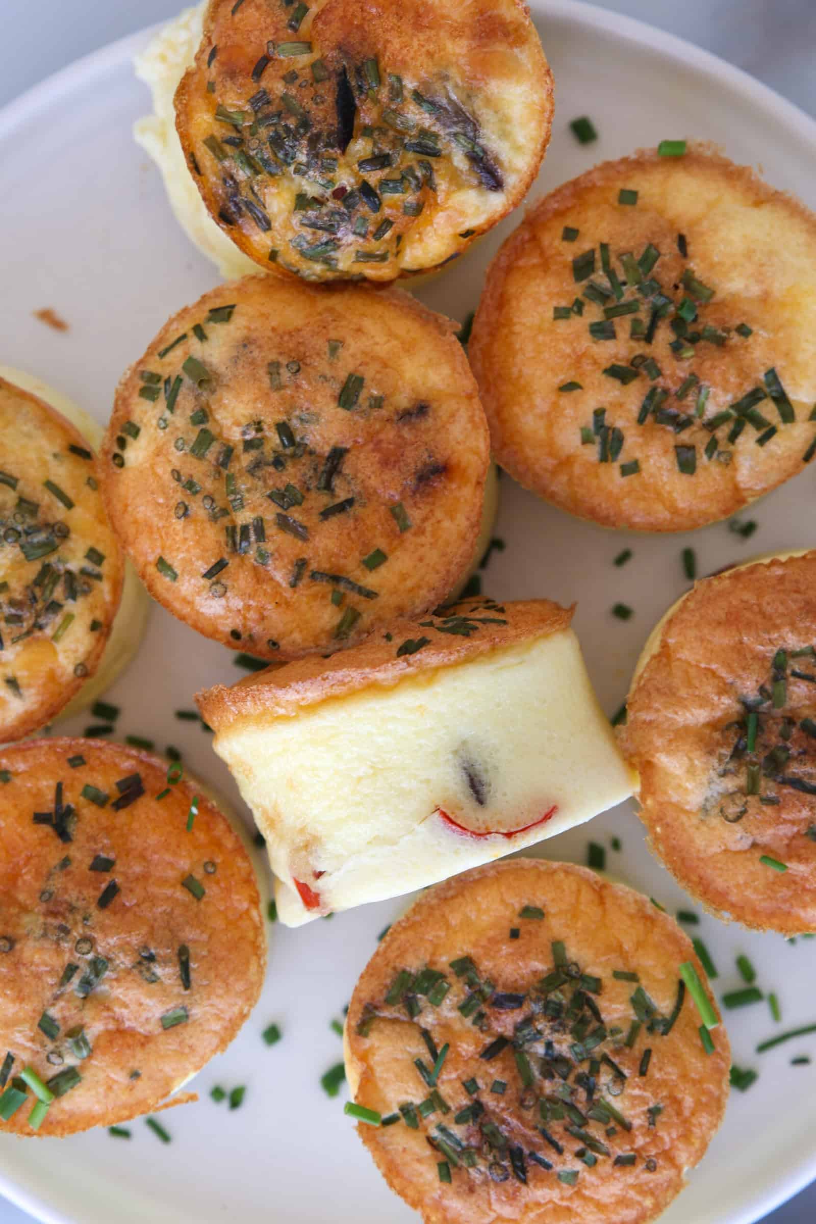 A close-up of a white plate filled with mini cottage cheese egg bites topped with chopped chives and visible vegetable pieces, arranged in a circular pattern.