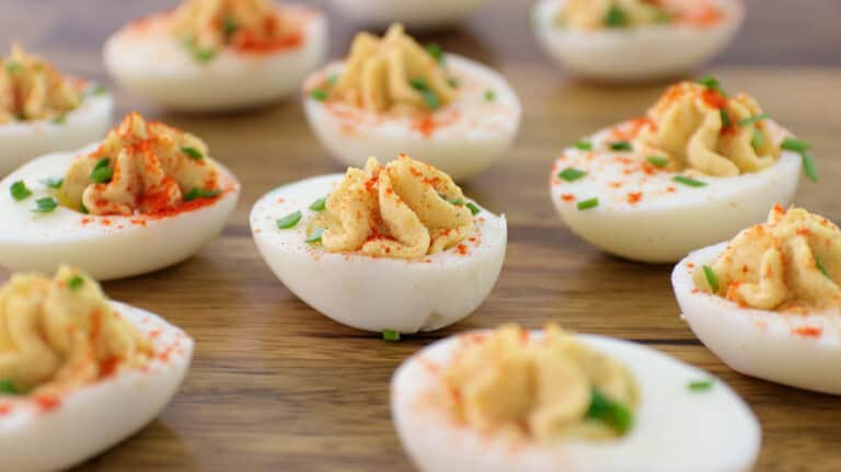 Several deviled eggs topped with chopped chives and sprinkled with paprika are arranged on a wooden surface. The eggs are halved with creamy, piped yolk filling in the center.