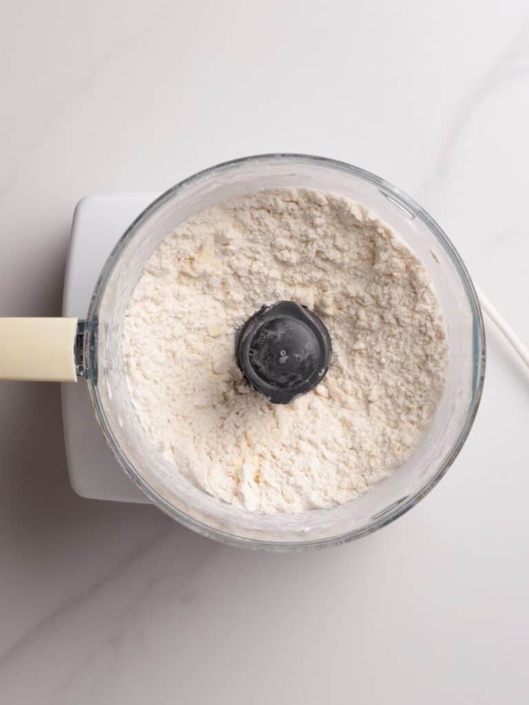 A food processor bowl filled with flour mixture sits on a white countertop, viewed from above. The food processor has a white base and a light-colored handle on the left side.