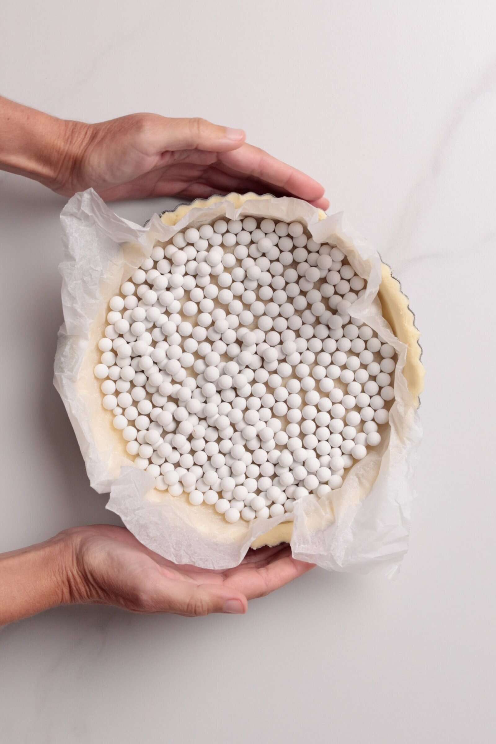 A pair of hands holds an unbaked pie crust lined with parchment paper and filled with white ceramic baking beads, ready for blind baking.