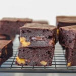 Two thick, square cottage cheese brownies with chunks of yellow nuts and gooey melted chocolate inside are stacked on a cooling rack, with more brownies in the background.