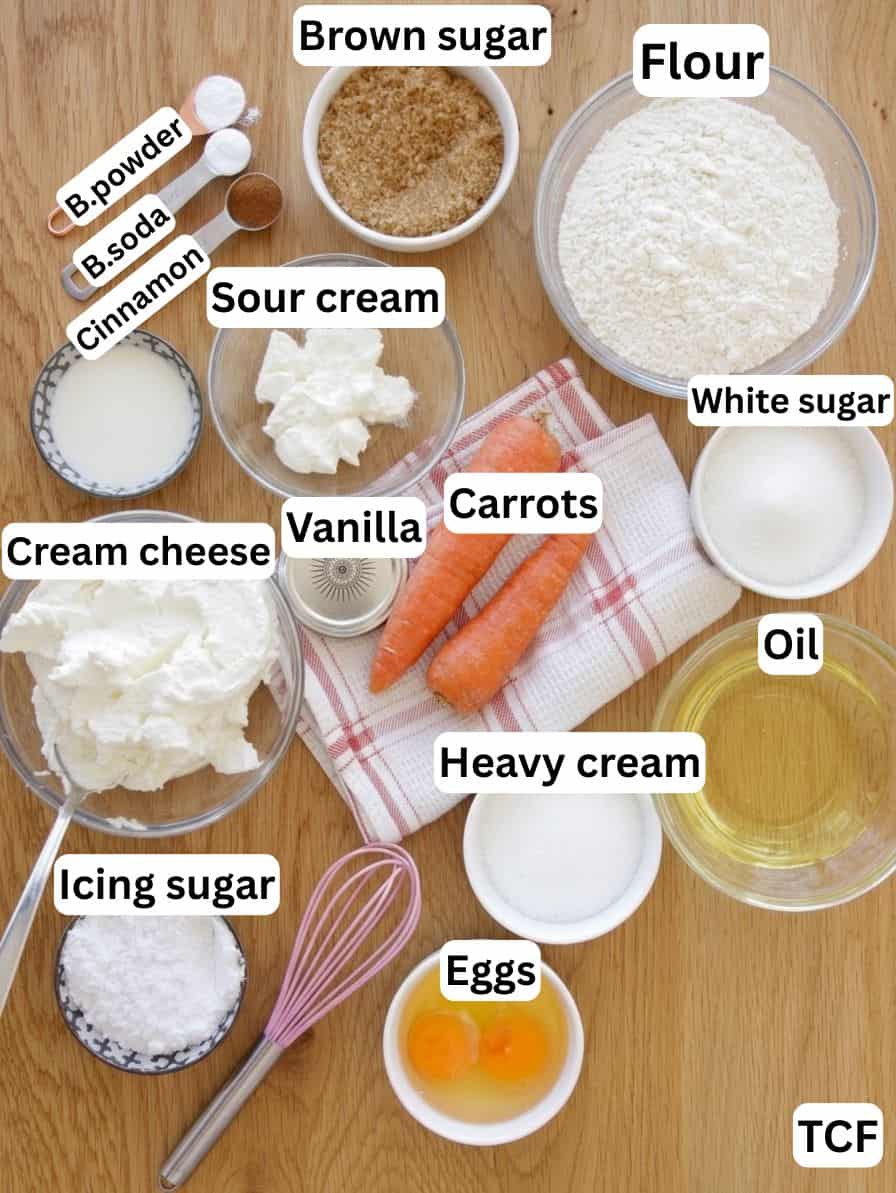 Top-down view of baking ingredients in bowls and plates, including flour, brown and white sugar, oil, eggs, carrots, cream cheese, heavy cream, sour cream, vanilla, icing sugar, baking powder, baking soda, and cinnamon.
