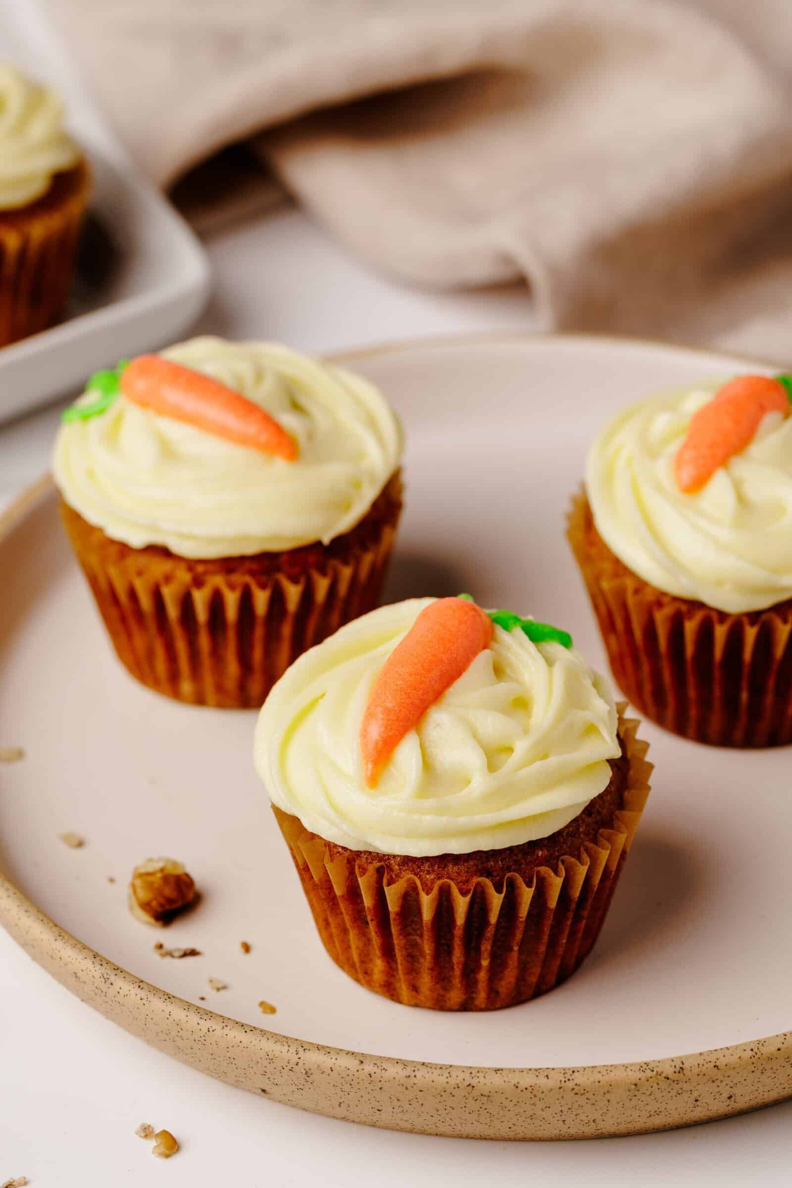 Three carrot cake cupcakes with cream cheese frosting and small carrot decorations on top, arranged on a beige plate with a few crumbs nearby. A beige cloth and more cupcakes are visible in the background.