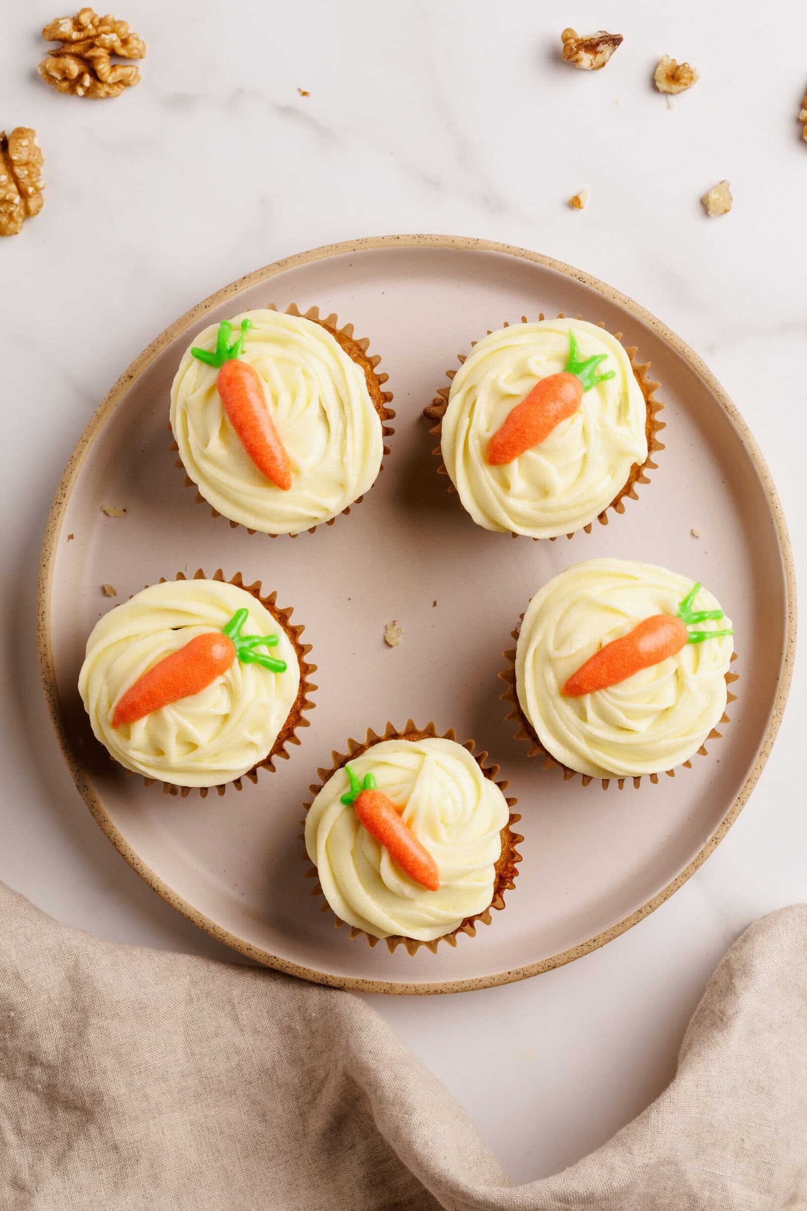 Five cupcakes with swirled white frosting and small carrot decorations on top, arranged on a round beige plate, with walnuts scattered nearby on a marble surface.