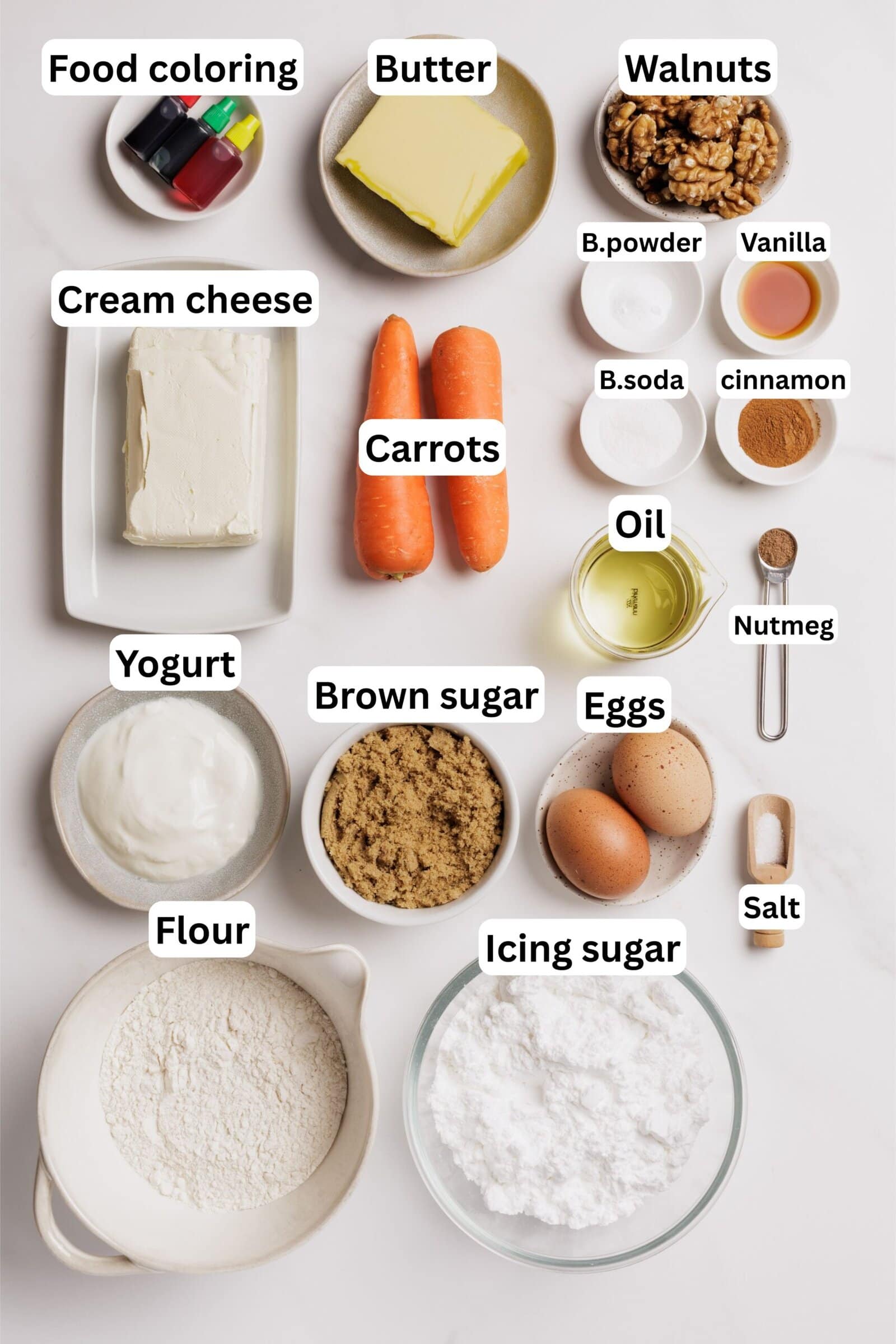 A flat lay of labeled baking ingredients, including food coloring, butter, walnuts, cream cheese, carrots, yogurt, brown sugar, flour, icing sugar, baking powder, vanilla, baking soda, cinnamon, oil, eggs, salt, and nutmeg.