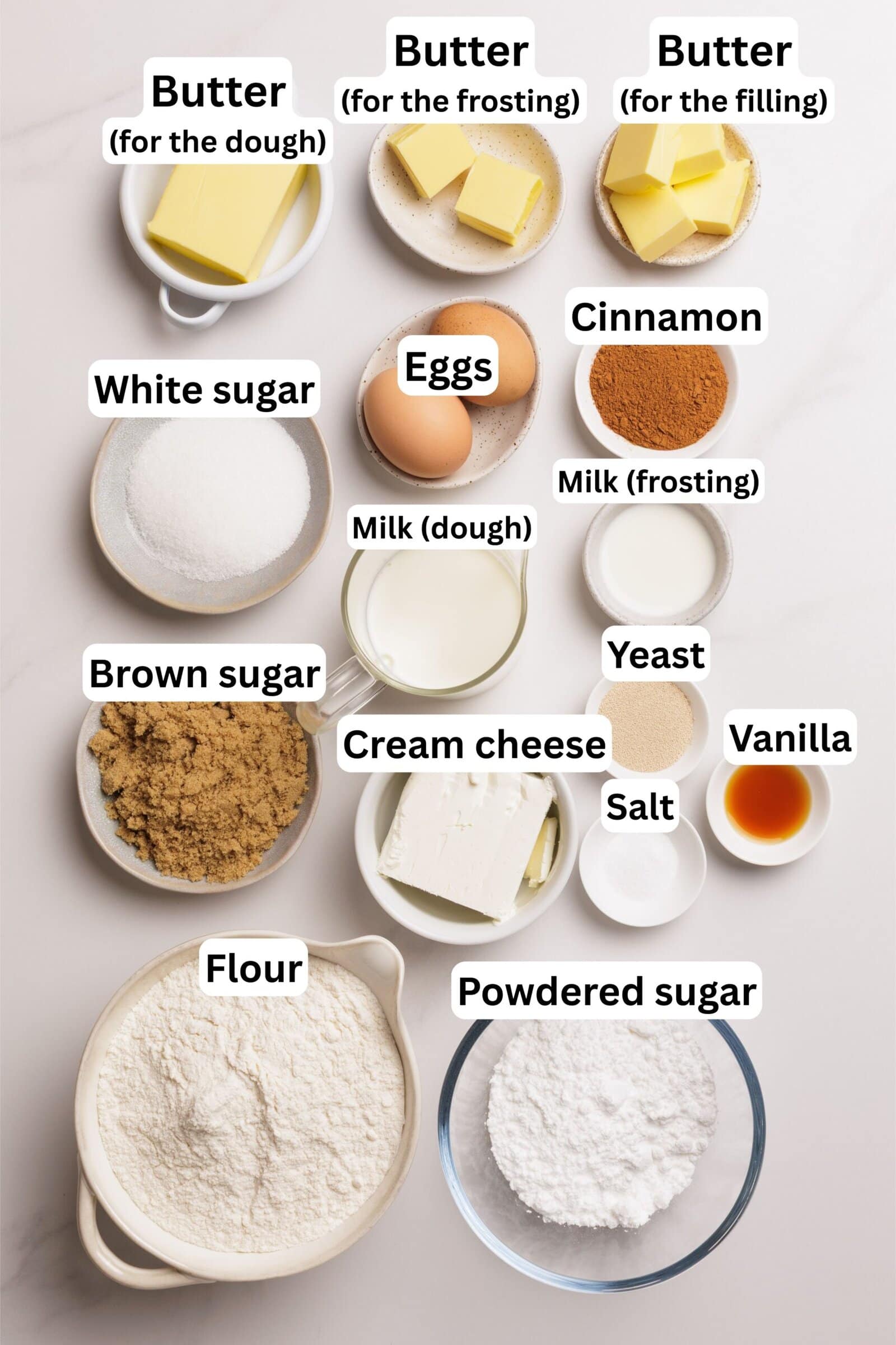 Overhead view of labeled baking ingredients in bowls: butter (for dough, frosting, and filling), white sugar, eggs, cinnamon, brown sugar, milk (for dough and frosting), yeast, cream cheese, salt, vanilla, flour, and powdered sugar.