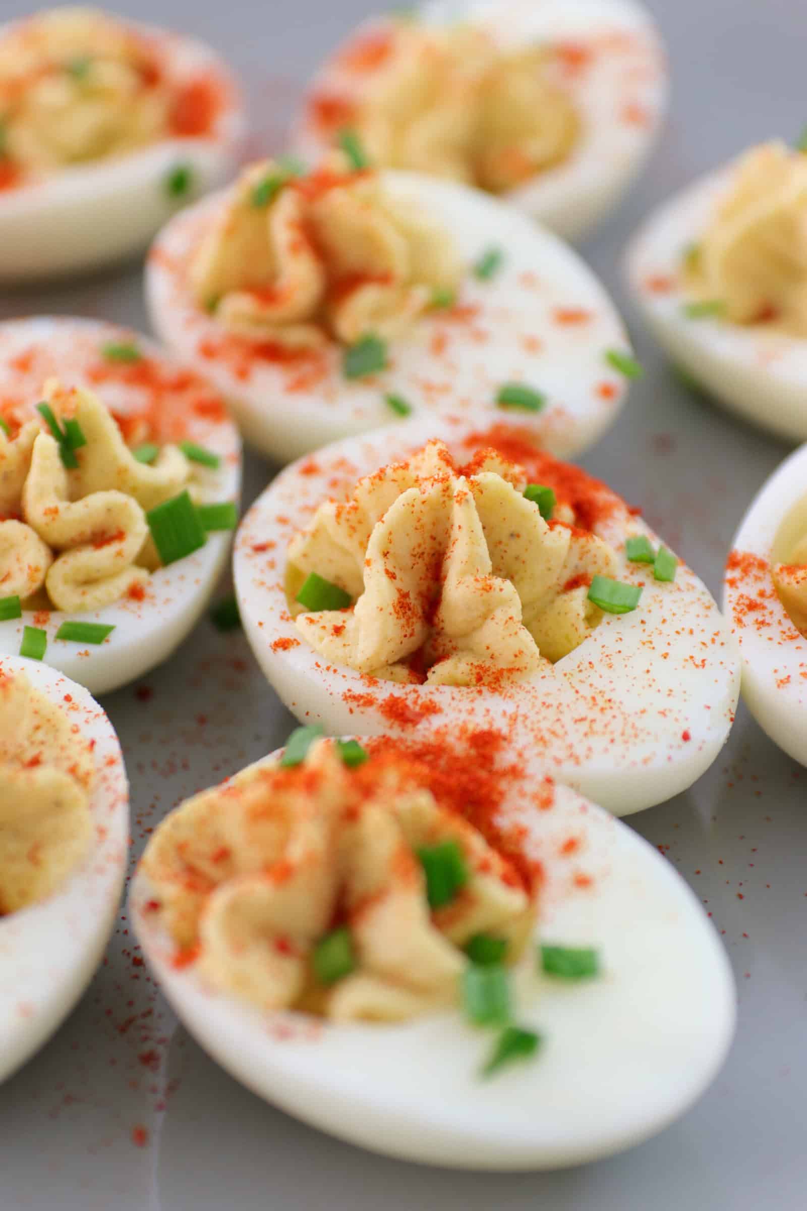Close-up of deviled eggs topped with paprika and chopped chives, arranged on a light-colored surface. The eggs are halved, with creamy yolk filling piped in the center.