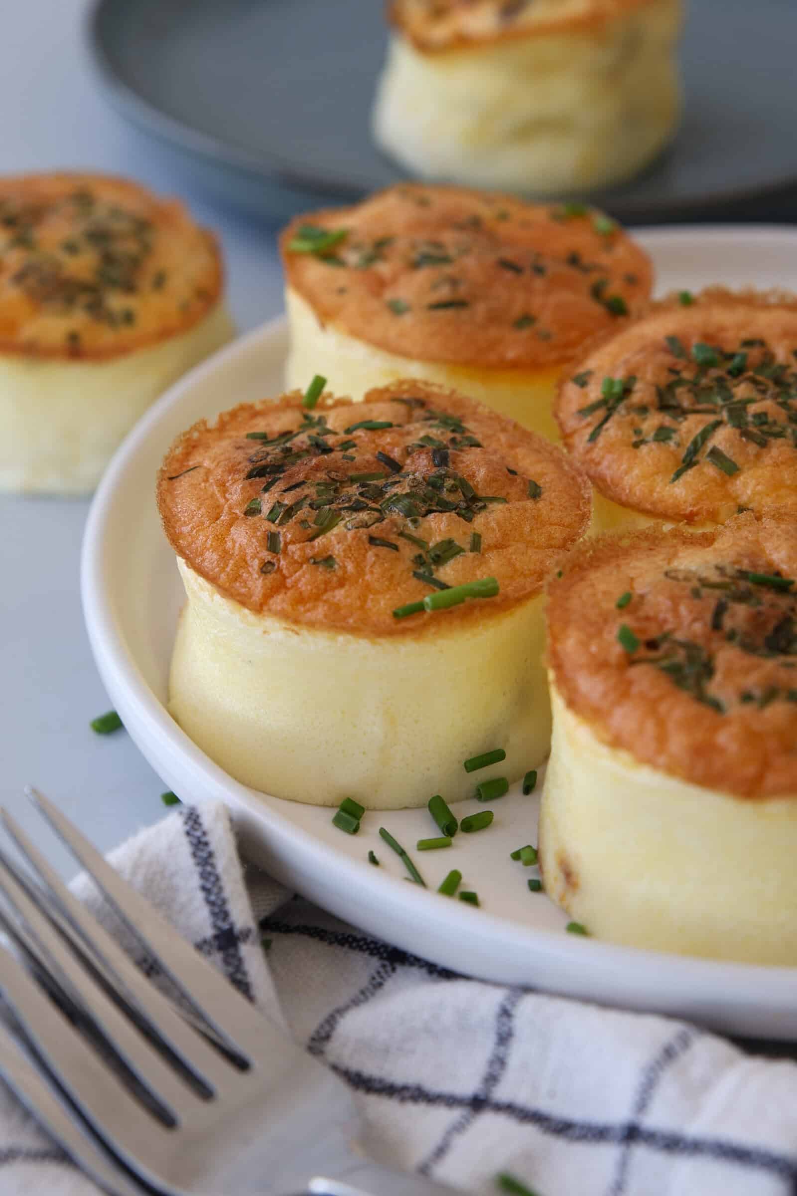 Three fluffy baked cottage cheese egg bites topped with chopped herbs are served on a white plate. A fork and a checkered napkin are in the foreground, with more soufflés visible in the background.