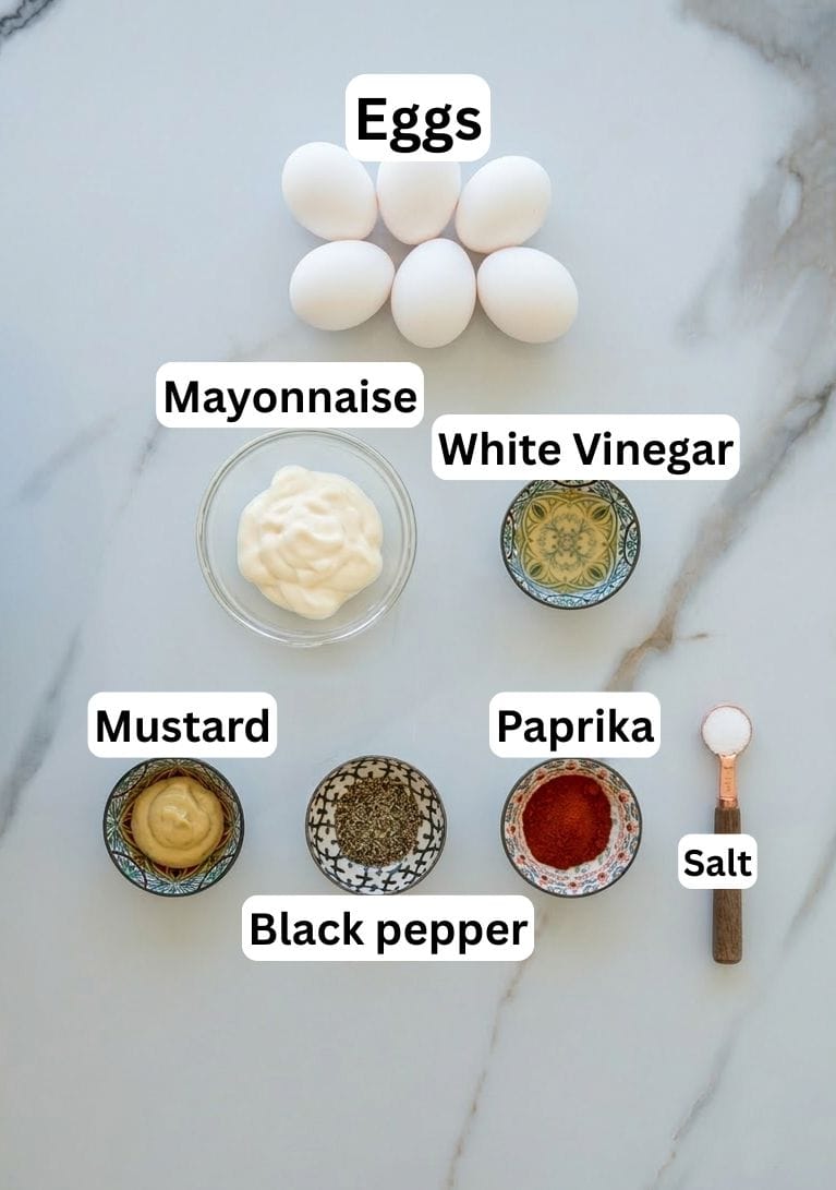 A flat lay of deviled egg ingredients: six eggs, a bowl of mayonnaise, a bowl of white vinegar, small bowls of mustard, black pepper, paprika, and a measuring spoon of salt, all labeled.