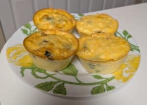 Four mini egg muffins with melted cheese and greens, placed on a white plate with a yellow floral design, sitting on a white countertop.