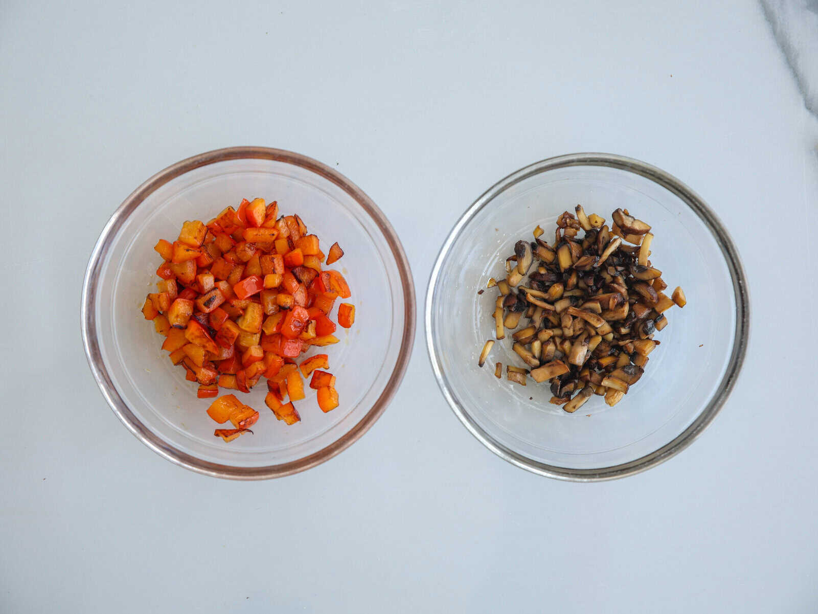 Two glass bowls on a white surface; the left bowl contains chopped red and orange bell peppers, and the right bowl contains chopped mushrooms.
