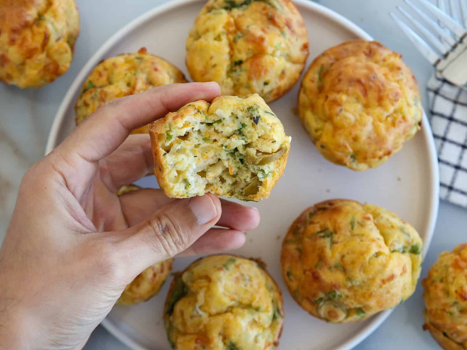 A hand holds a savory muffin with a bite taken out, showing a soft, herby interior. More muffins rest on a round white plate in the background, along with a fork and checkered napkin.