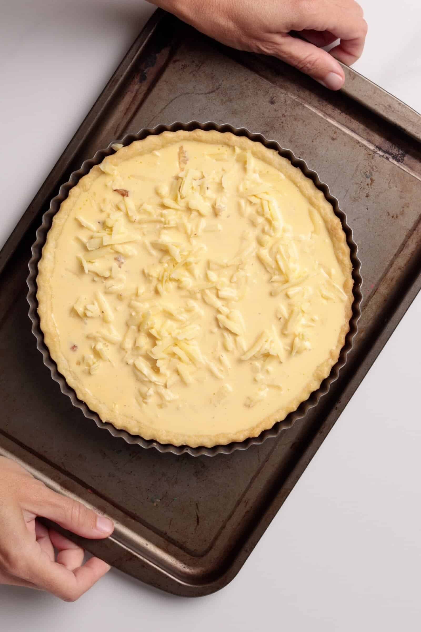 A person places an unbaked quiche topped with grated cheese on a round tart tin onto a baking tray, with both hands visible holding the tray.