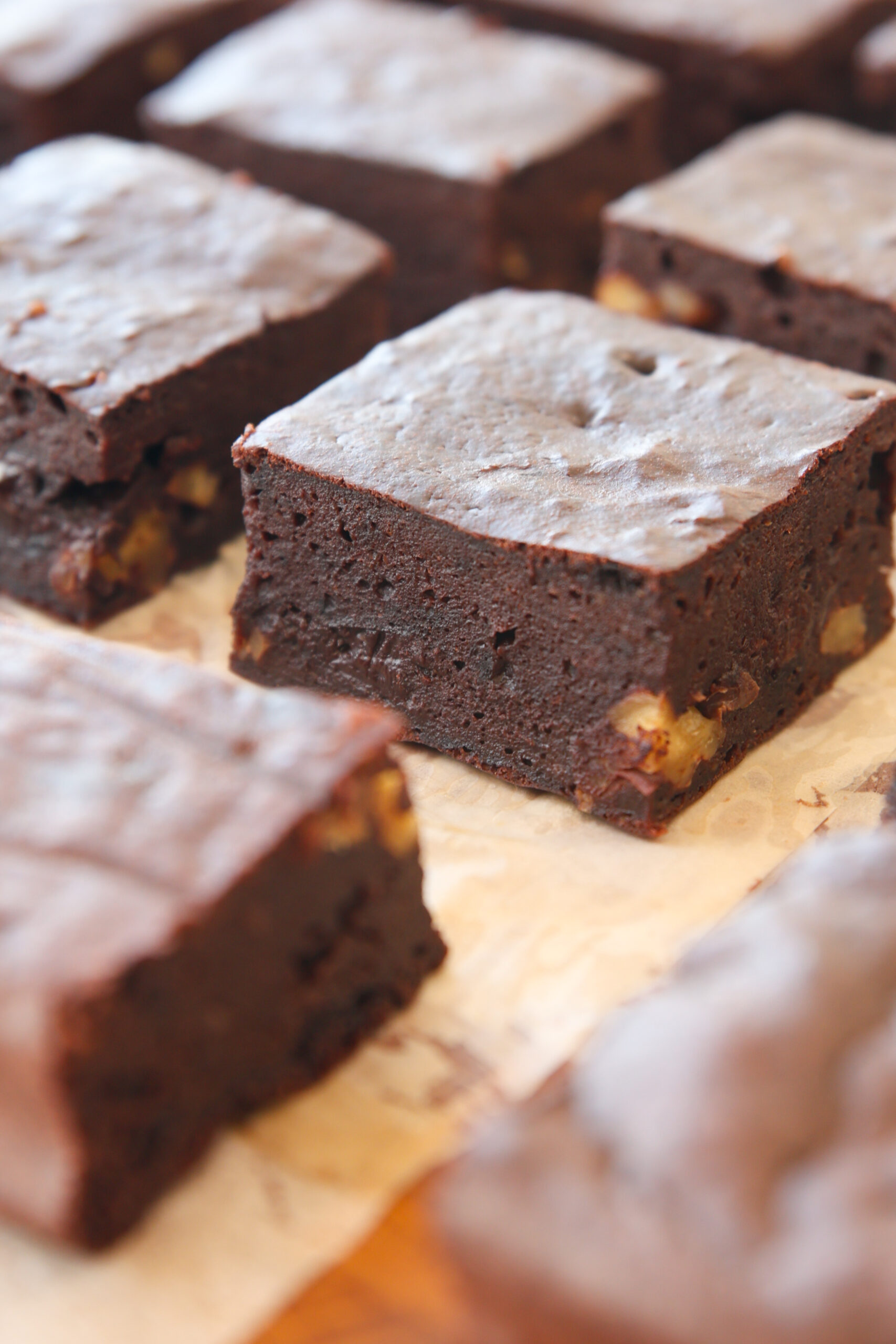 Close-up of thick, fudgy high-protein chocolate brownies with visible walnut pieces, arranged in rows on parchment paper. The brownies have a rich, moist texture and a slightly glossy top.