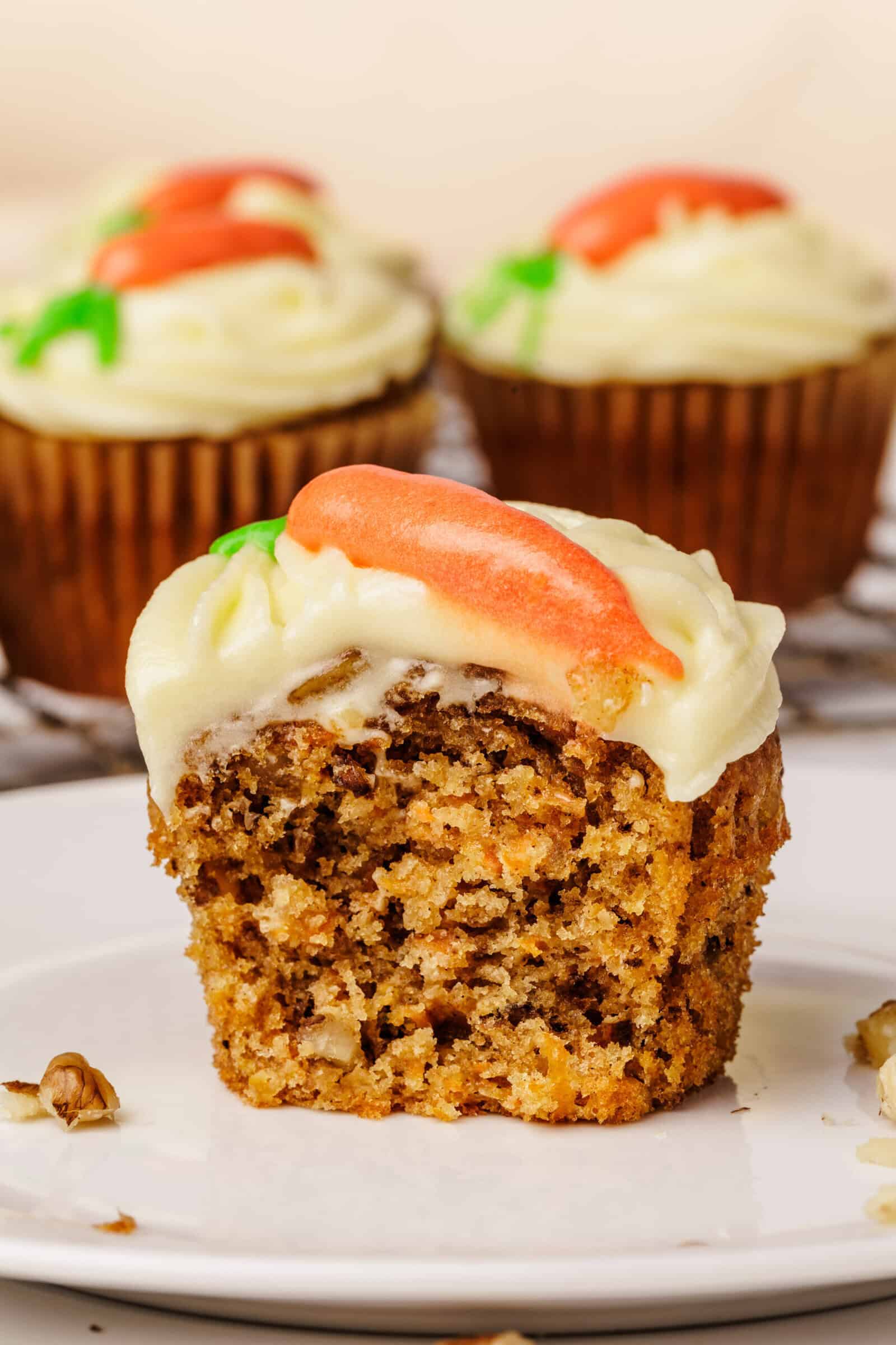 A carrot cake cupcake with cream cheese frosting and a small icing carrot on top, cut in half to show its moist, nutty interior. Two more decorated cupcakes are blurred in the background.