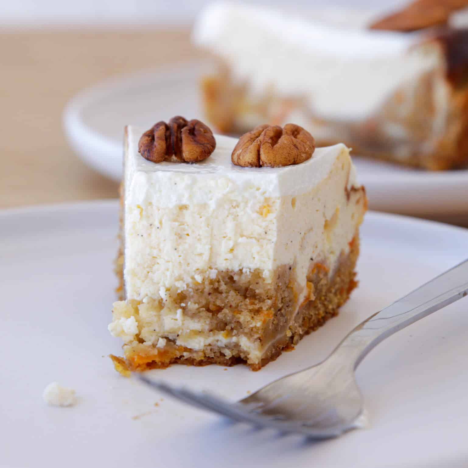 A slice of creamy carrot cake cheesecake with a crumbly base and two pecans on top sits on a white plate next to a fork, with another slice visible in the background.