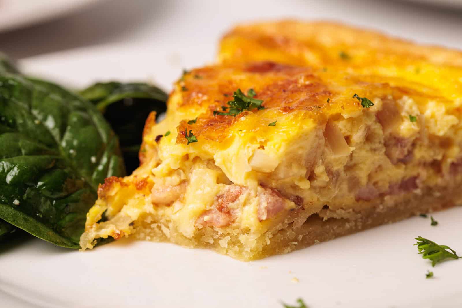 A close-up of a slice of quiche with a golden, cheesy top and visible pieces of ham, served on a white plate next to a small portion of fresh spinach leaves.