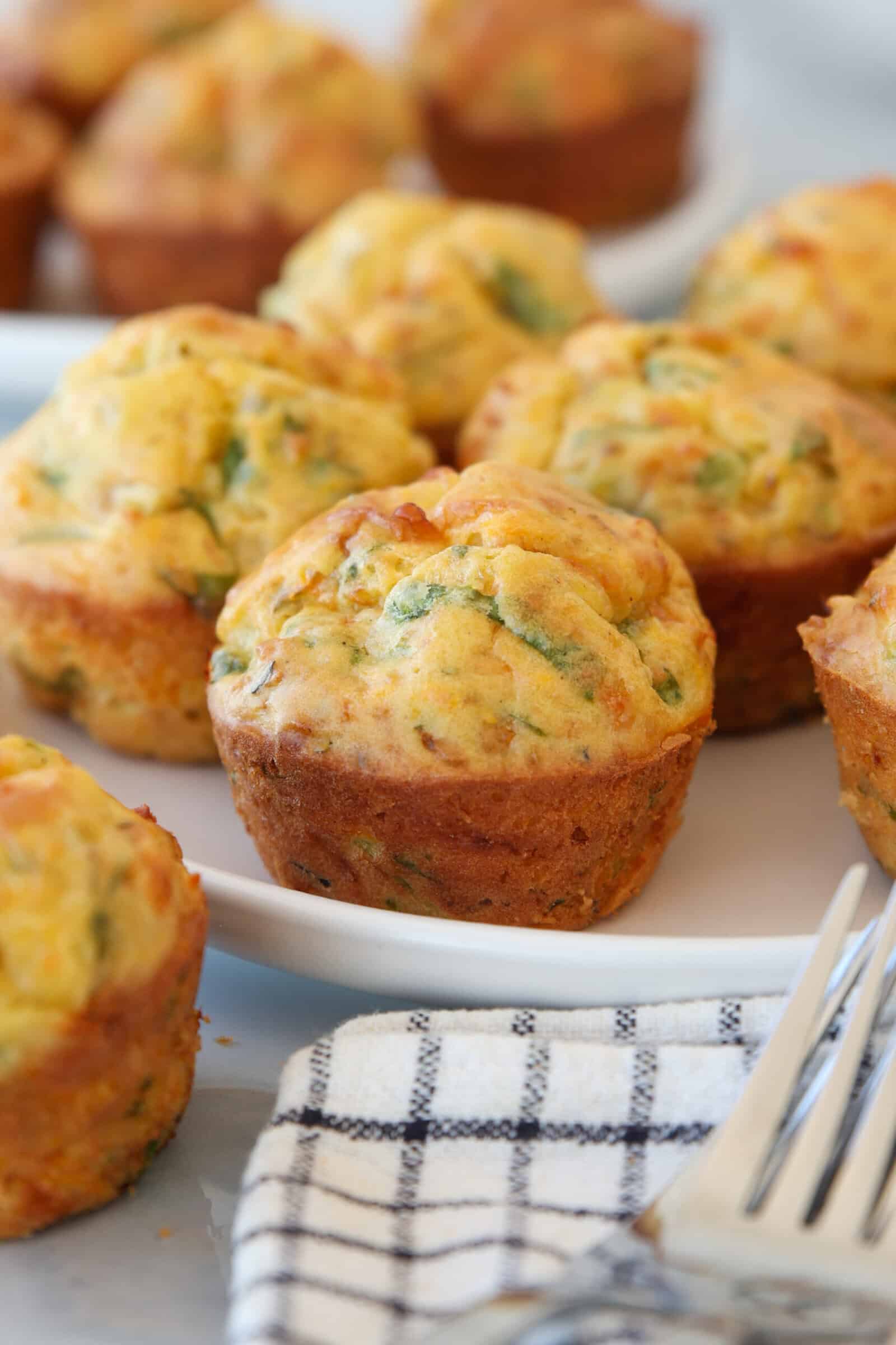 Golden brown savory muffins with visible herbs are arranged on a white plate, with additional muffins in the background. A checkered cloth and a fork are placed in the foreground.