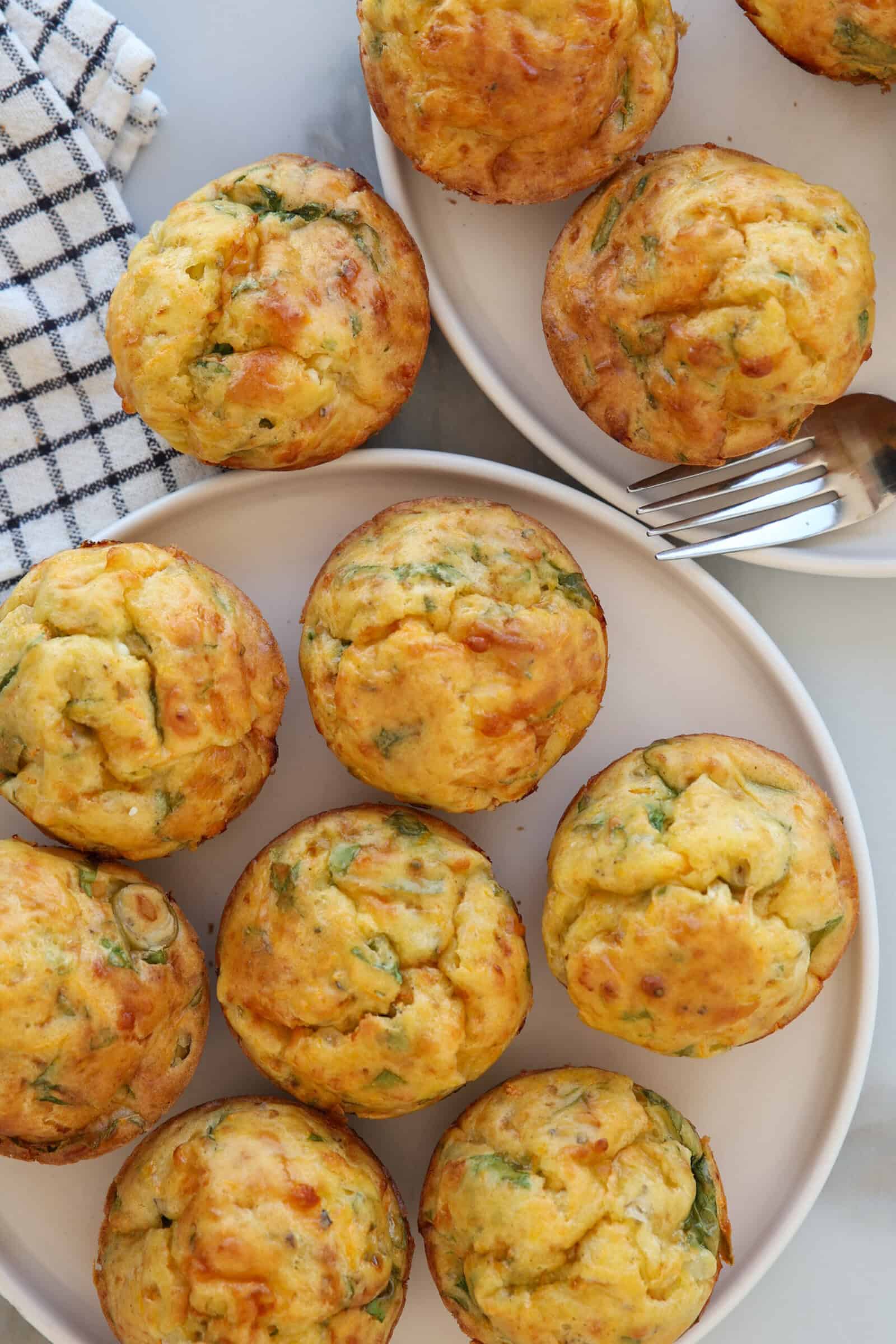 Golden, fluffy cottage cheese muffins with green herbs are arranged on two white plates; a fork and a checkered kitchen towel are nearby on a marble surface. The muffins appear savory and freshly baked.