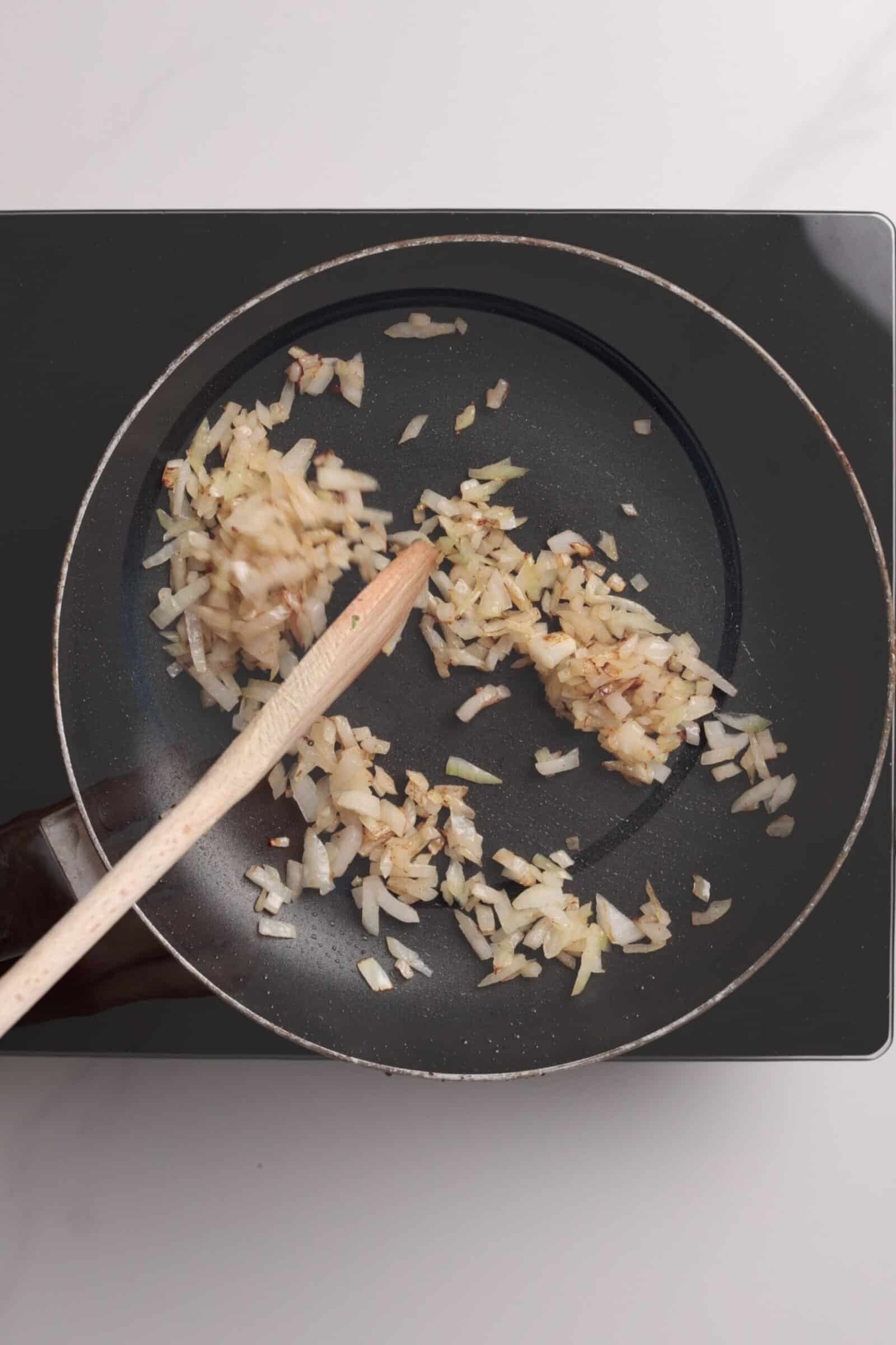 Chopped onions being sautéed in a black frying pan on a stovetop, stirred with a wooden spoon.