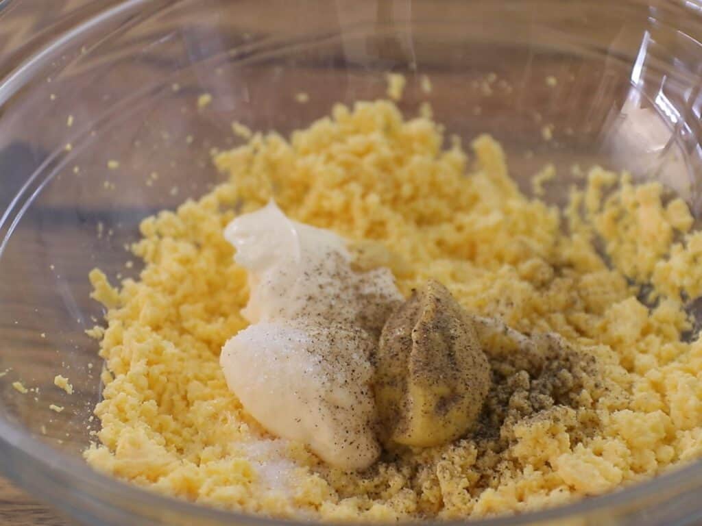 Crumbled cooked egg yolks in a glass bowl topped with dollops of mayonnaise, mustard, ground black pepper, and a pinch of salt, ready to be mixed.