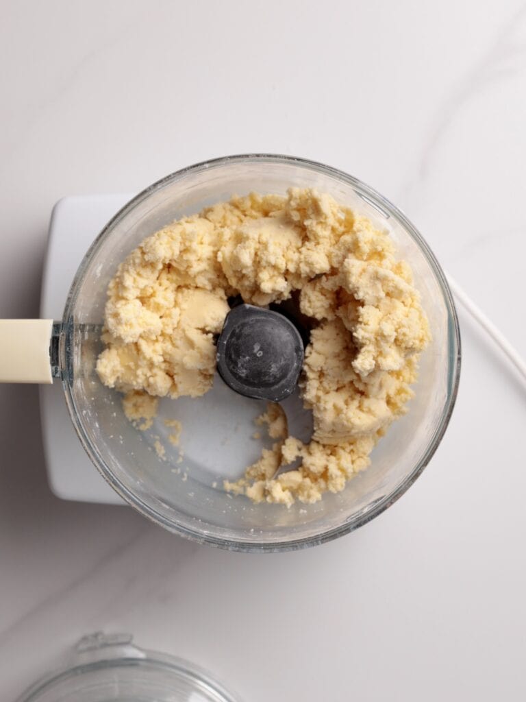 A food processor with a pale, crumbly dough mixture inside sits on a white countertop with part of its lid visible nearby.