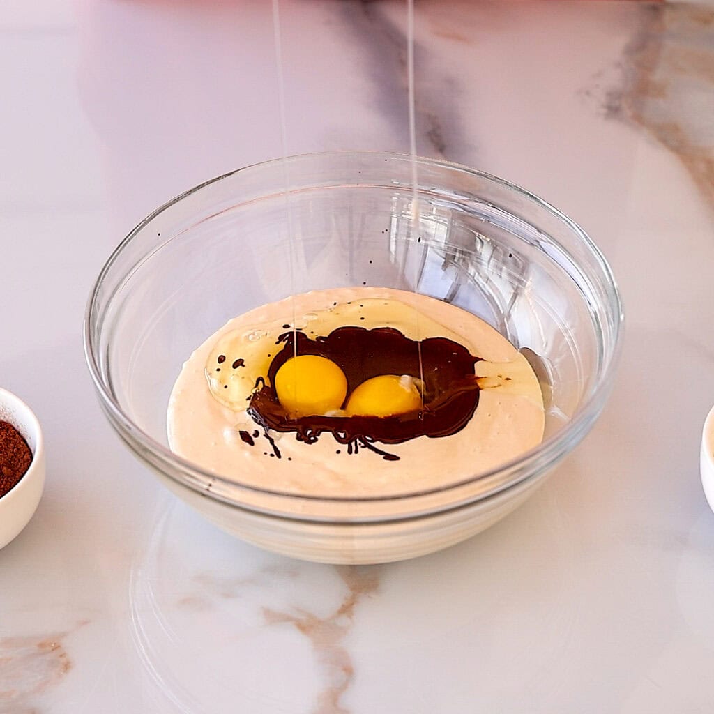 A glass bowl with flour, two cracked eggs, and chocolate sauce on a marble countertop, surrounded by small bowls of ingredients.
