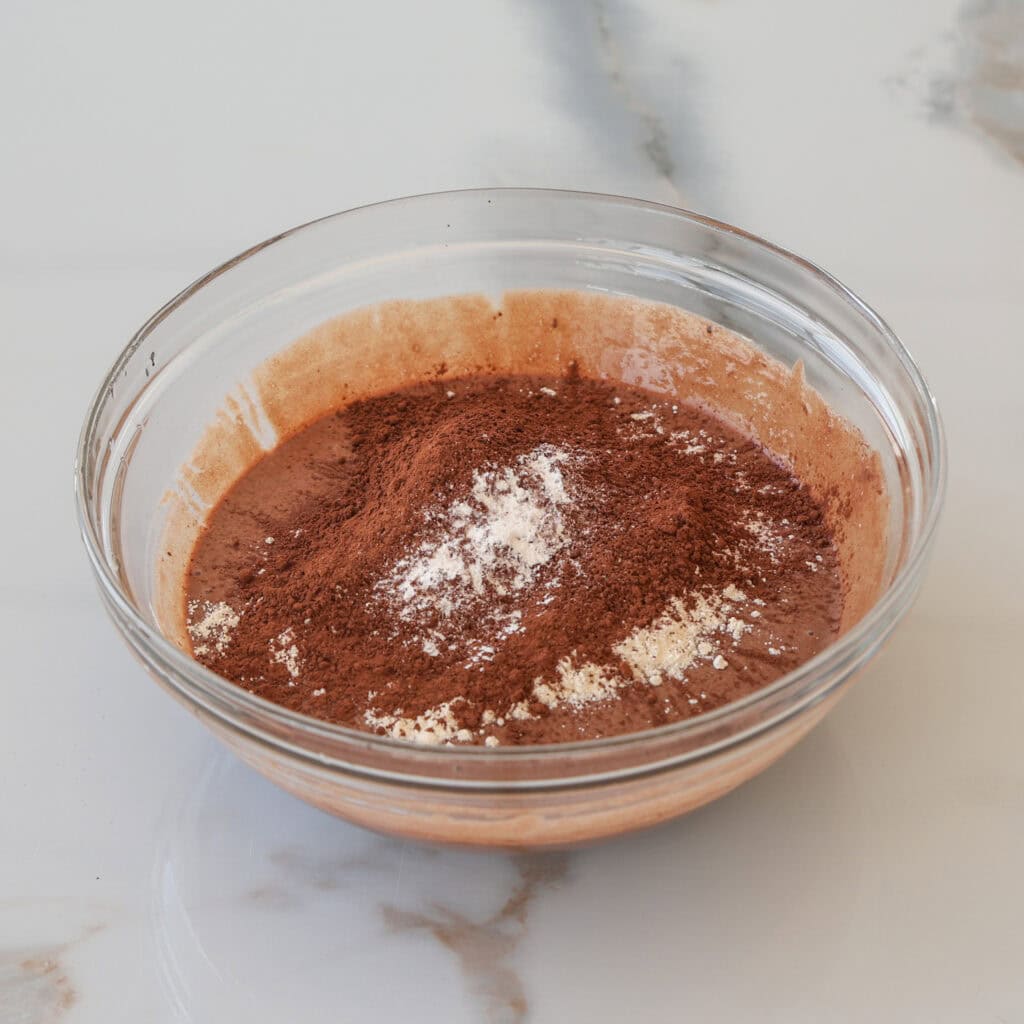A clear glass bowl filled with chocolate cake batter and a mound of cocoa powder and flour on top, sitting on a white marble surface.
