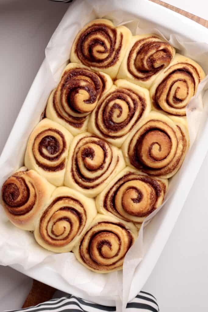 A close-up of a baking dish filled with neatly arranged, golden-brown cinnamon rolls with a visible cinnamon swirl pattern, resting on white parchment paper.