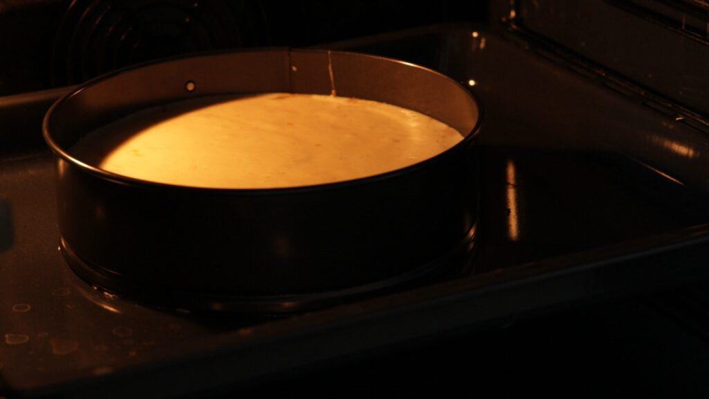 A round cake pan filled with batter is placed in an oven, baking. Warm light from the oven illuminates the pan and batter, which has risen slightly. The oven door is open, and the metal racks are visible.