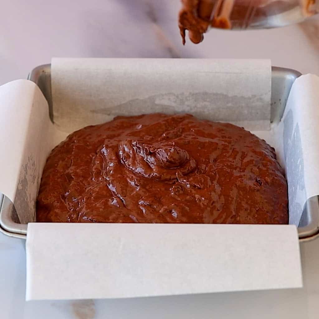A square baking pan lined with parchment paper is filled with chocolate brownie batter. Some batter is being poured into the center, creating a mound. The pan sits on a marble countertop.
