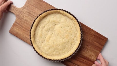 A baked homemade quiche crust with fork marks sits in a round tart pan on a wooden cutting board, held at each end by two hands against a light background.