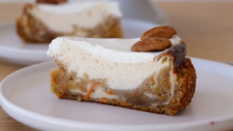 A slice of carrot cake cheesecake with creamy white frosting, topped with two pecan halves, sits on a white plate. Another slice of cake is blurred in the background.