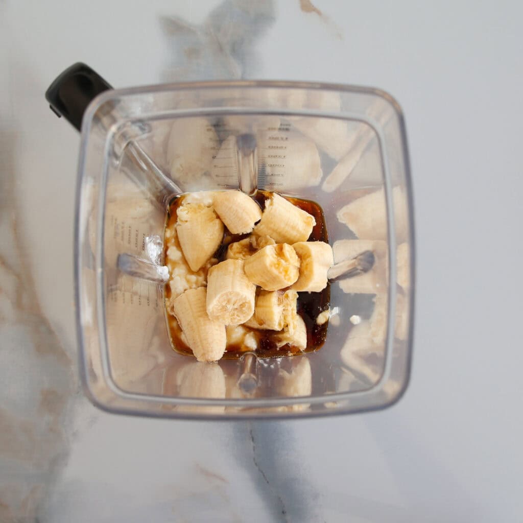 Overhead view of a blender containing chopped bananas, a brown liquid (possibly syrup), and a white substance, all ready to be blended on a marble countertop.