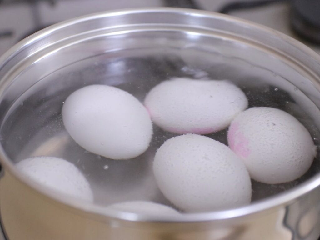 Six white eggs are being boiled in a stainless steel pot filled with water on a stovetop. Steam and bubbles are visible, indicating the water is hot.