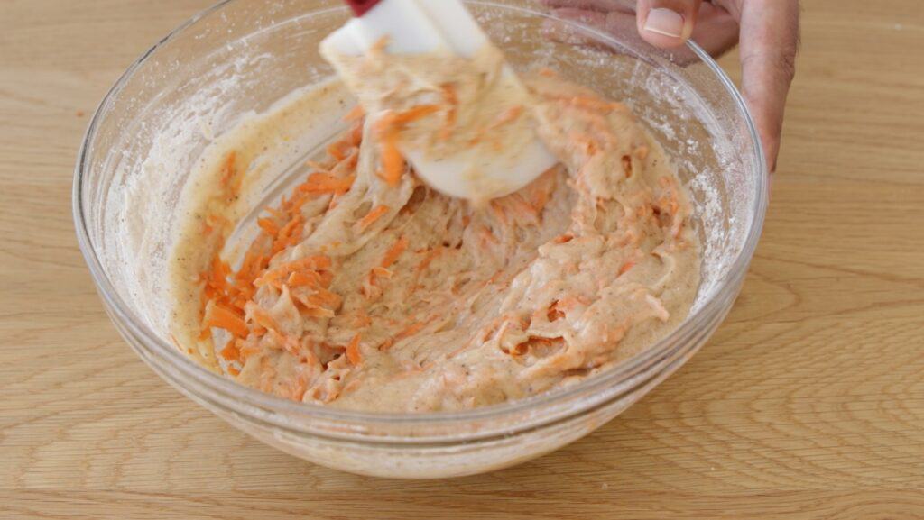 A hand stirs carrot cake batter with a spatula in a clear glass bowl on a wooden surface. Shredded carrots are visible in the thick mixture.