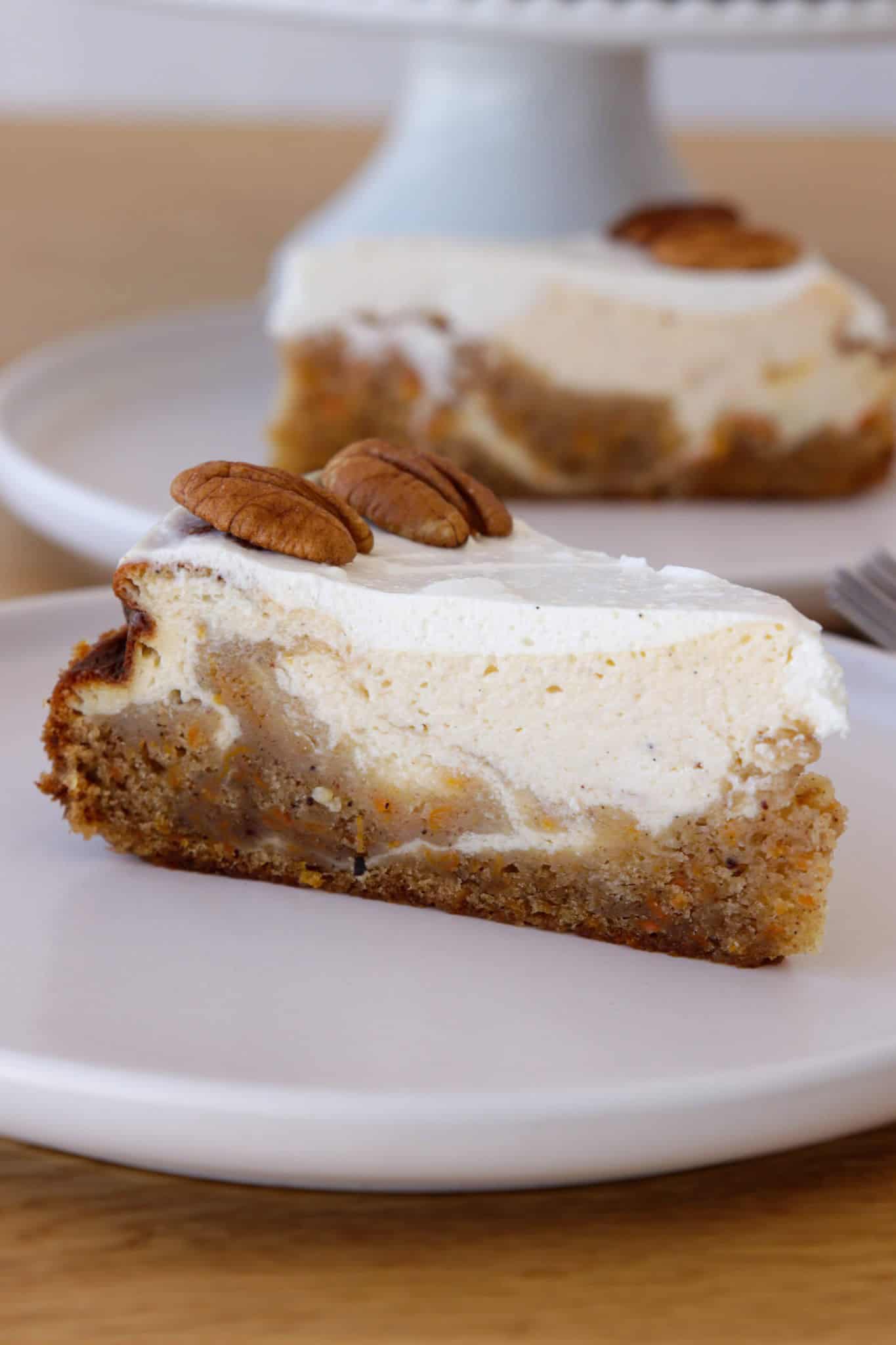 A slice of a carrot cake cheesecake, and sour cream topping, garnished with whole pecans, served on a white plate with another slice in the background.