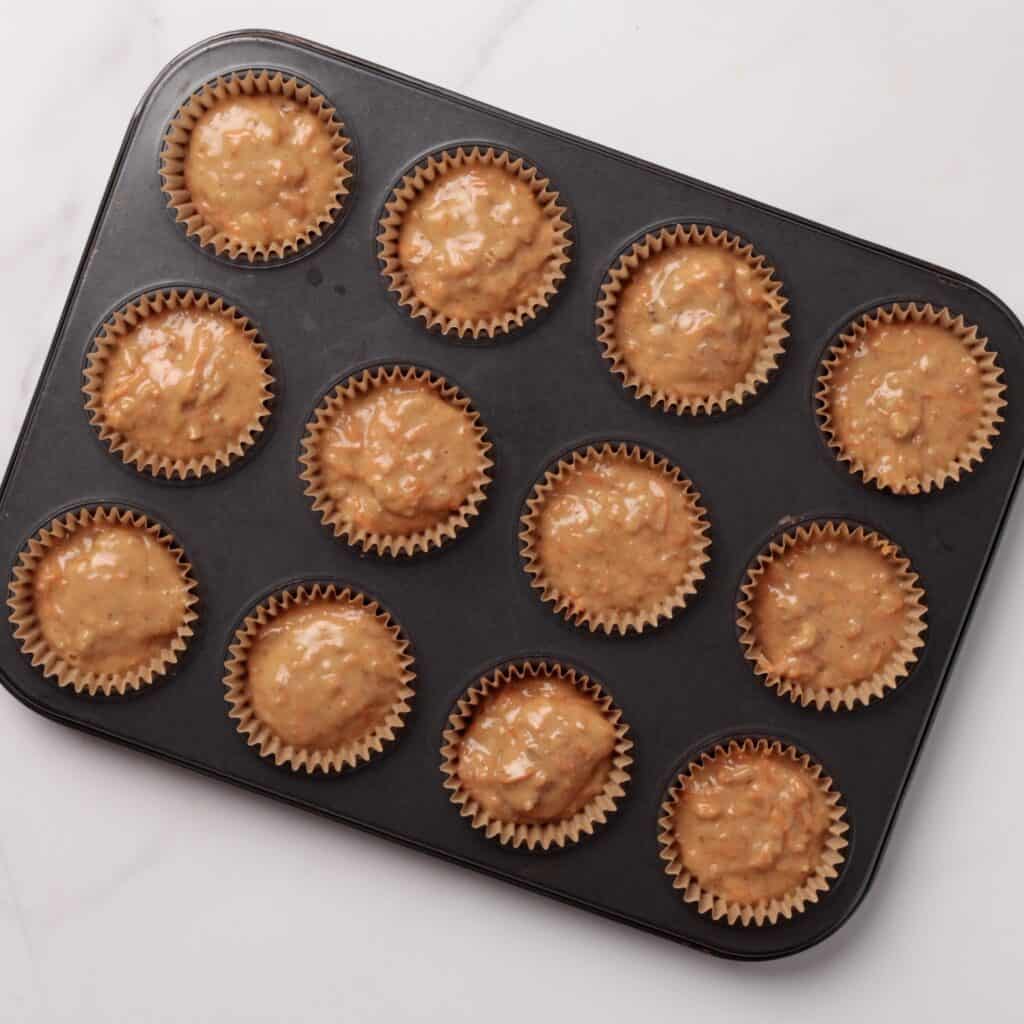 A muffin tin filled with twelve paper liners, each containing unbaked muffin batter with visible pieces of nuts or fruit, sits on a white marble surface.