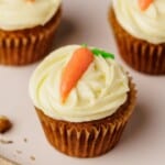 A carrot cupcake with creamy white frosting and a small carrot decoration on top, displayed on a light surface with two more cupcakes in the background.