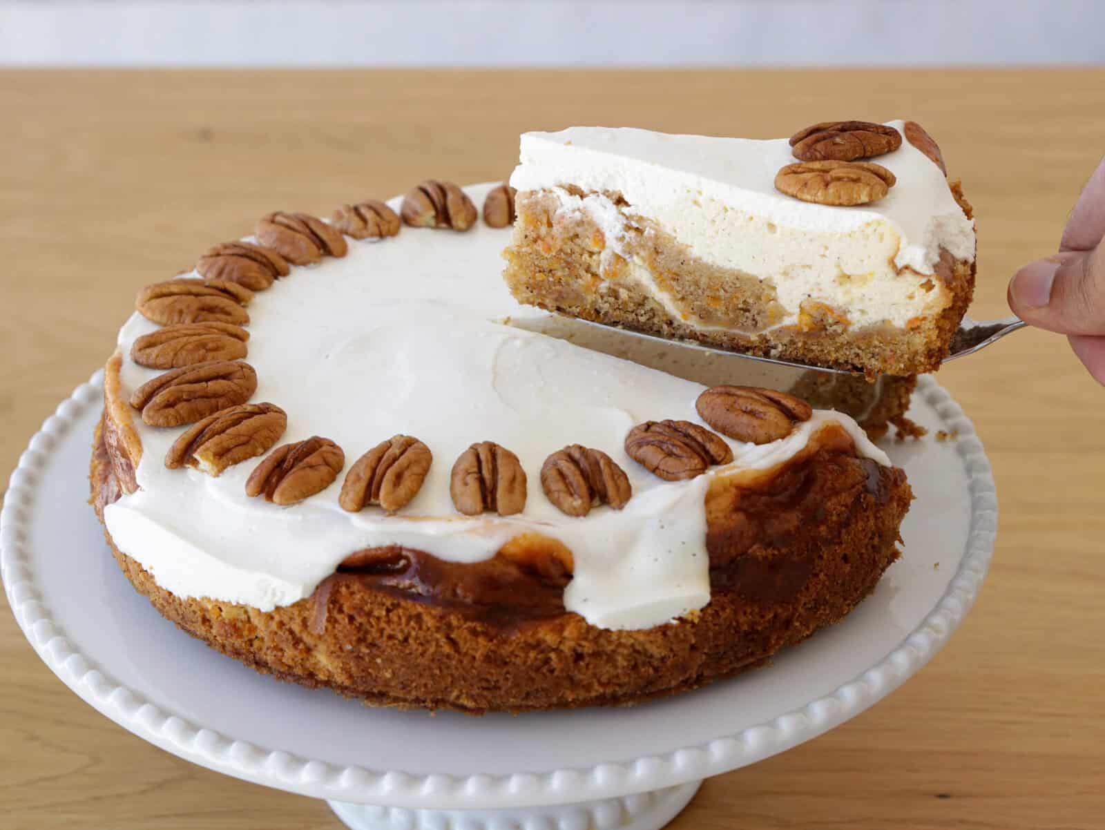 A hand lifts a slice of creamy cheesecake topped with pecans from a whole cheesecake, which is decorated with whipped cream and whole pecans, sitting on a white cake stand.