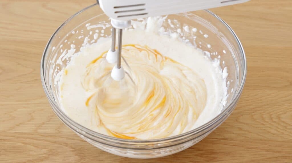 A hand mixer blending a white creamy mixture with streaks of yellow in a clear glass bowl on a wooden surface.