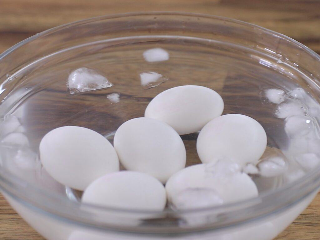 Six hard-boiled eggs are cooling in a glass bowl filled with ice water and ice cubes, placed on a wooden surface.