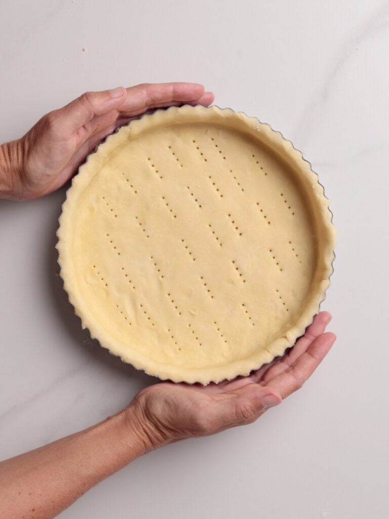 Two hands are holding a round tart pan filled with unbaked pie crust, which has small fork holes evenly pricked across its surface. The background is a light-colored countertop.