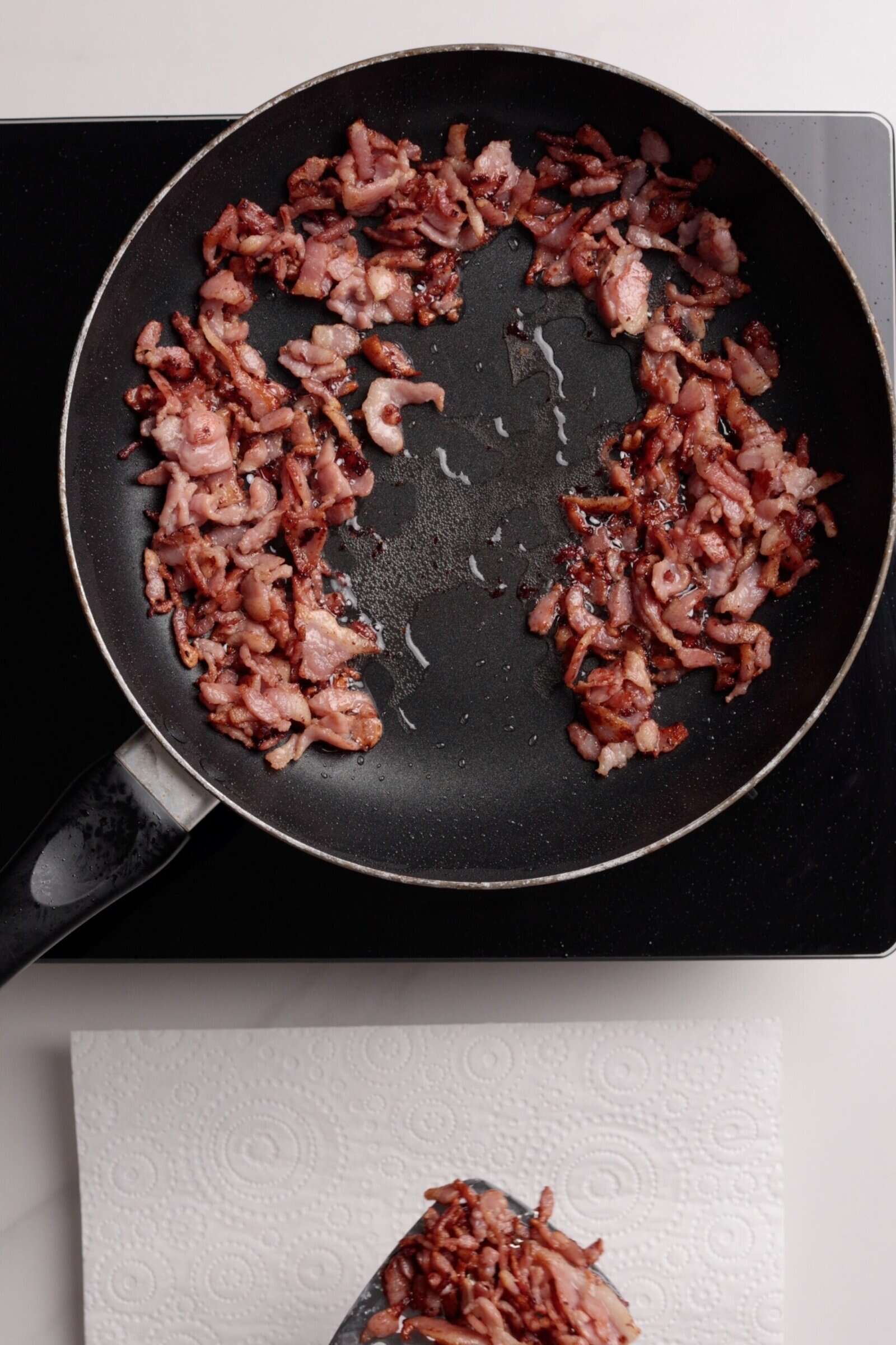 A frying pan with cooked bacon pieces arranged in a ring sits on a stovetop, while some bacon is being placed onto a paper towel below to drain excess oil.