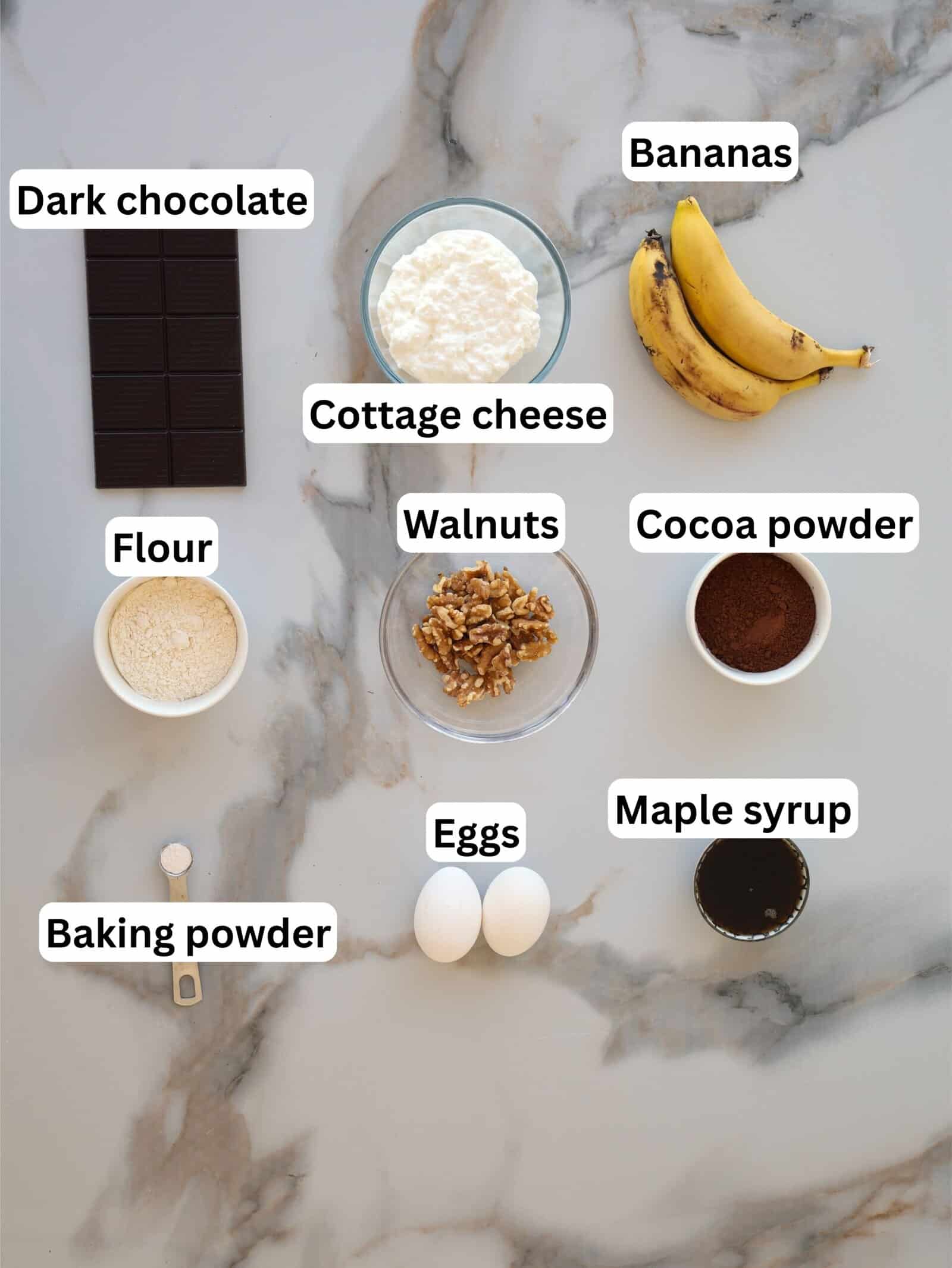 Ingredients for baking are laid out on a marble surface with labels: dark chocolate, cottage cheese, bananas, flour, walnuts, cocoa powder, baking powder, eggs, and maple syrup.