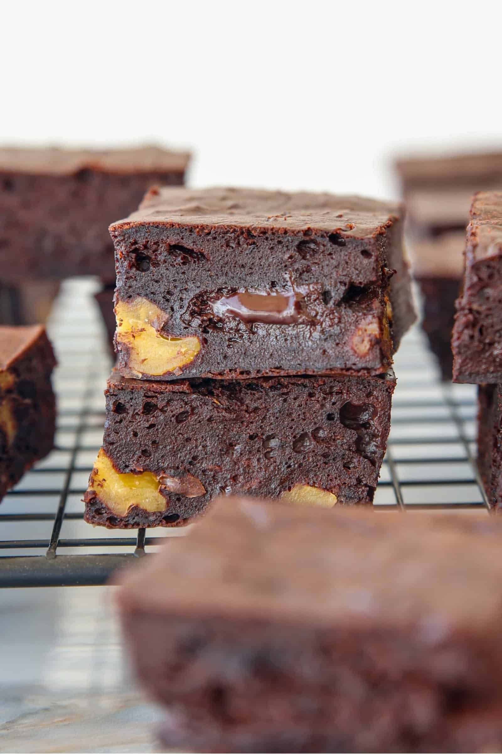 Two thick, fudgy cottage cheese brownies with visible chocolate chunks and pieces of yellow fruit are stacked on a wire cooling rack, with more brownies blurred in the background.