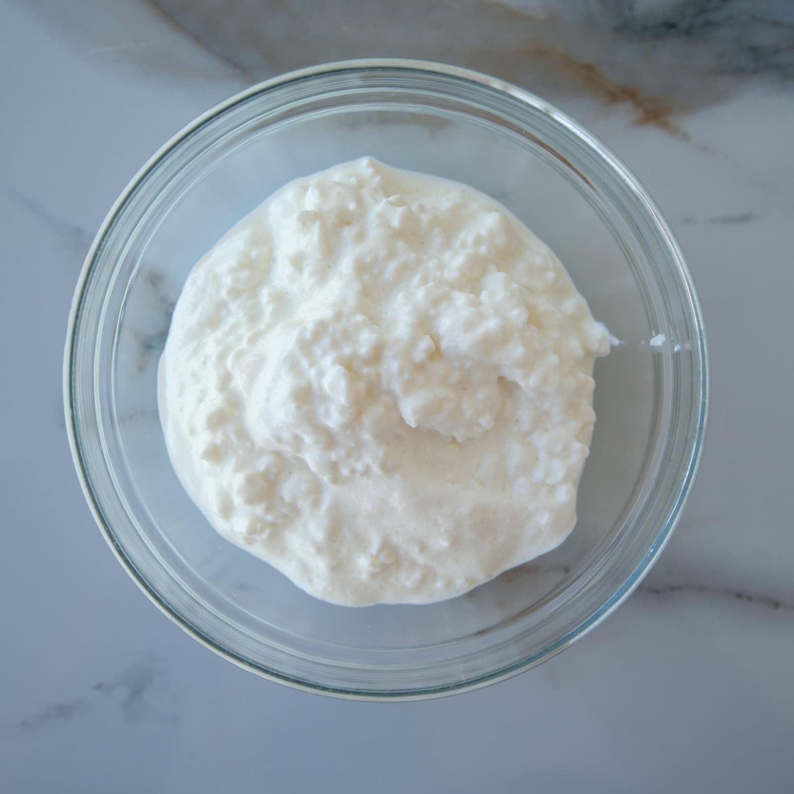 A clear glass bowl filled with white cottage cheese sits on a light marble surface.