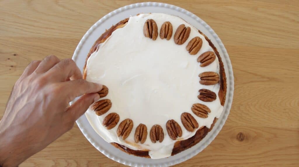 A hand places a pecan half onto a round cake with white frosting, already decorated with a ring of pecans around the edge, on a white plate and wooden surface.
