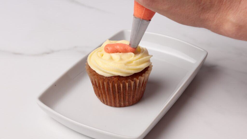 A hand decorates a cupcake with white frosting using a piping bag with orange icing, on a rectangular white plate and white marble surface.