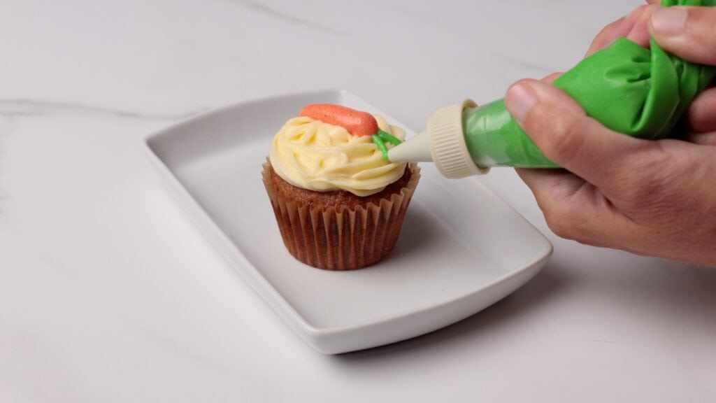 A hand decorates a cupcake with green icing on a white rectangular plate, adding details next to a small orange carrot made of frosting on top of creamy yellow swirl frosting.
